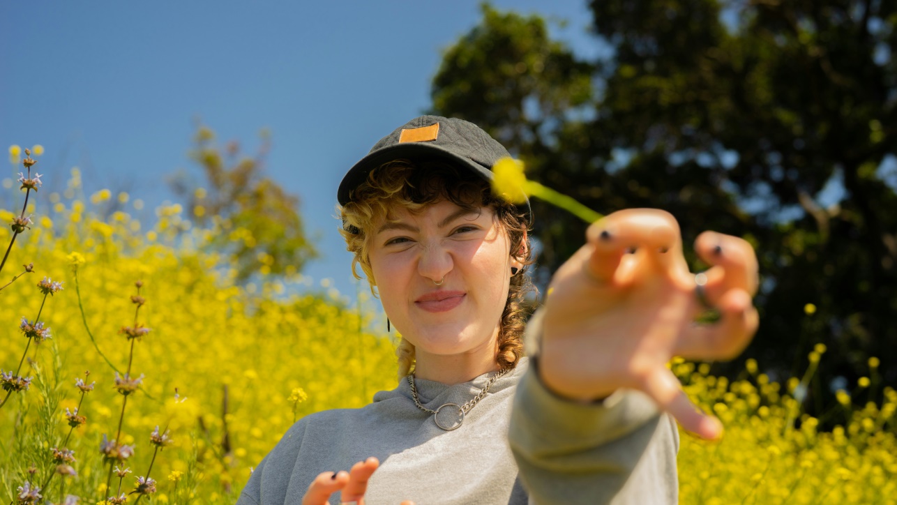 Portræt af en person i et felt af vilde blomster, der griner med øjnene let sammenknebne, mens hånden strækkes tæt mod kameraet. Den varme, solfyldte natur og kroppens spontane gestus formidler energi, kreativitet og nærvær, som kan spejle den proces, terapi med ACT understøtter – særligt for dig, der lever med ADHD eller er i tvivl om udredning og søger støtte til at finde ro, retning, balance og kontakt til egne værdier.