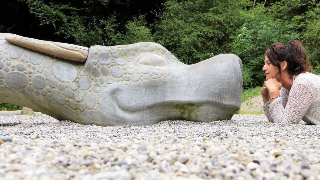 En person ligger i grus og møder en stor skulptur af et smilende dragehoved i et roligt, grønt område – et billede på nysgerrighed, kontakt og tilstedeværelse.